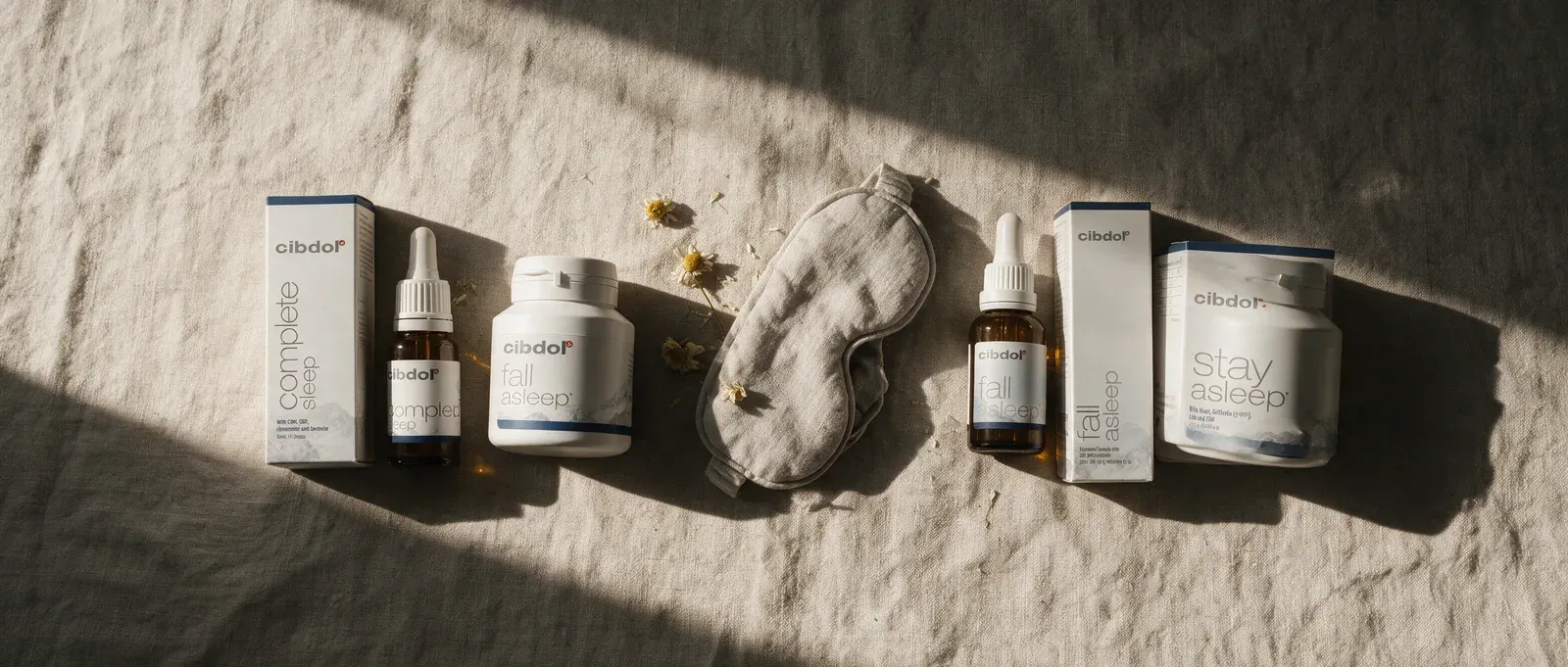 Cibdol's four sleep products, Complete Sleep, Fall Asleep Capsules, Fall Asleep Liquid and Stay Asleep Capsules, laid out on linen with chamomile and a sleep mask